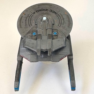 Puede incluir: Maqueta gris de una nave espacial con el texto "NCC-31060-A" en la parte superior. El modelo tiene una secci&oacute;n superior circular, una secci&oacute;n media rectangular y dos extensiones largas y rectangulares con detalles azules. El modelo tiene detalles azules y rojos.