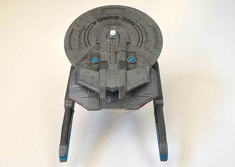 Puede incluir: Maqueta gris de una nave espacial con el texto "NCC-31060-A" en la parte superior. El modelo tiene una secci&oacute;n superior circular, una secci&oacute;n media rectangular y dos extensiones largas y rectangulares con detalles azules. El modelo tiene detalles azules y rojos.