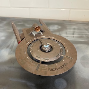 Puede incluir: Modelo detallado de una nave espacial, con un casco principal circular y dos góndolas de curvatura. La nave es principalmente marrón con detalles en rojo y azul. El texto "NCC-1279" es visible en el casco.