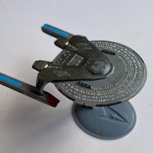 Könnte beinhalten: Graues Modell eines Star Trek-Raumschiffs mit kreisförmigem Hauptrumpf und zwei Warpgondeln mit blauen Akzenten. Das Modell ist auf einem grauen Ständer mit dem Starfleet-Abzeichen montiert.