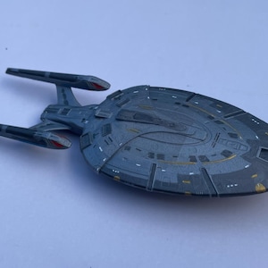 Puede incluir: Maqueta gris de una nave espacial con un casco principal en forma de platillo y dos góndolas de curvatura. El modelo tiene detalles rojos y amarillos y está sobre un fondo blanco. La nave espacial es una réplica de la USS Enterprise.