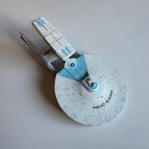 Puede incluir: Maqueta de una nave estelar de Star Trek, con una sección de platillo blanca con el texto "NCC-5386" y detalles azules. La nave tiene un casco rectangular y dos góndolas de curvatura.
