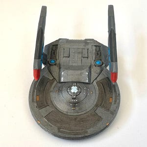 Puede incluir: Maqueta gris de nave espacial con cuerpo circular y dos alas verticales. Las alas tienen detalles en rojo y azul. El cuerpo de la nave presenta paneles detallados y un elemento central azul brillante.