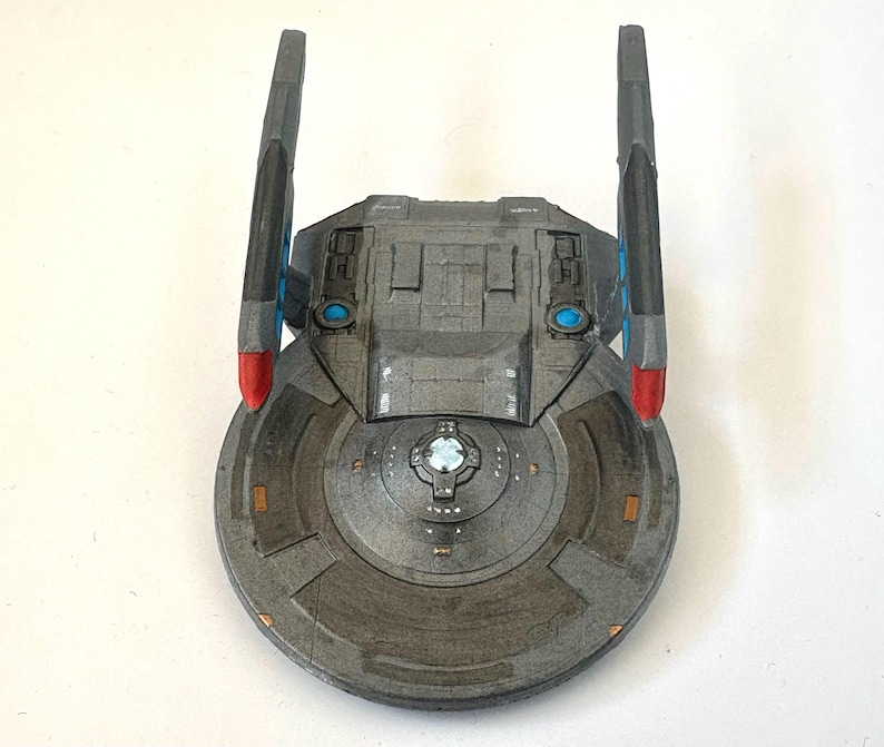 Puede incluir: Maqueta gris de nave espacial con cuerpo circular y dos alas verticales. Las alas tienen detalles en rojo y azul. El cuerpo de la nave presenta paneles detallados y un elemento central azul brillante.