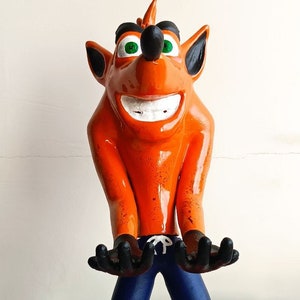 Può includere: Figurina di Crash Bandicoot arancione brillante, con un ampio sorriso, occhi verdi e dettagli neri. Indossa pantaloni blu, scarpe rosse con strisce bianche e poggia su una base circolare arancione. Braccia tese.