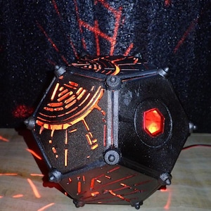 May include: A black, geometric lamp with a red light shining through the intricate cutouts. The lamp is shaped like a dodecahedron and has a hexagonal opening on one side.