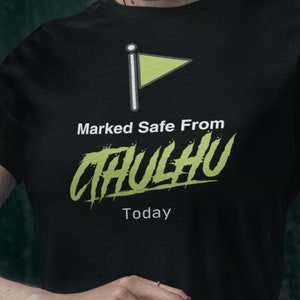 Puede incluir: Una mujer con una camiseta negra con una bandera verde y el texto "Marked Safe From Cthulhu Today". Está sosteniendo una bola de cristal en sus manos.