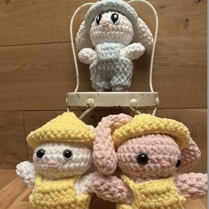 May include: Three crocheted bunny plushies wearing yellow overalls. The bunny on the left is white with a yellow hat, the bunny in the middle is light blue with a light blue hat, and the bunny on the right is pink with a yellow hat.