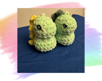 May include: Two plush, light green dinosaur toys with black eyes and a textured, fuzzy appearance. One has a yellow tail, and the other has green pom-poms on its head. They are sitting on a blue surface.
