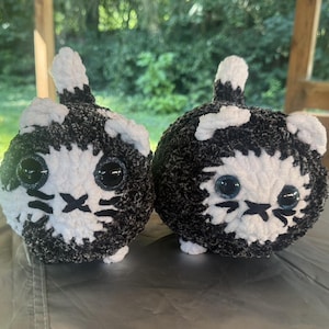 May include: Two handmade, round cat plushies. Each plushie is black and white, with large, round, black eyes and a stitched-on mouth and whiskers. The plushies are sitting on a gray surface.