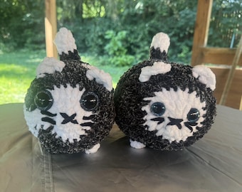 May include: Two handmade, round cat plushies. Each plushie is black and white, with large, round, black eyes and a stitched-on mouth and whiskers. The plushies are sitting on a gray surface.