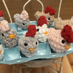 May include: A collection of nine crocheted chicks in a blue egg carton. The chicks are made with various colors of yarn, including white, gray, pink, and red.