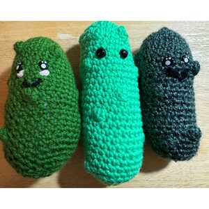 May include: Three handmade crochet pickle toys in varying shades of green and gray. Each pickle has a smiling face with black eyes and a stitched mouth. The pickles are displayed on a light brown wooden surface.