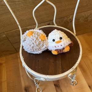 May include: Two crocheted stuffed animals, one white with brown and orange accents and the other brown and white, sitting on a white metal chair with a wooden seat.