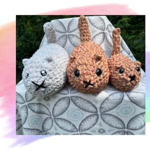 May include: Three crocheted cat plushies, one gray, one brown with orange accents, and one brown. The plushies are sitting on a gray and white patterned blanket.