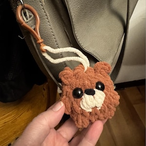 May include: A brown, plush teddy bear keychain with black eyes and a white muzzle. The bear is attached to a brown plastic clip with a white cord. The keychain is hanging from a gray bag.
