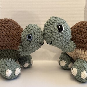 May include: Two handmade crochet turtles. The turtles have brown shells, green bodies, and white accents on their feet. They have large, round eyes and are positioned facing each other. The turtles are made of soft, chunky yarn.