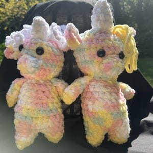 May include: Two handmade crochet unicorn dolls. The unicorns are made with soft, pastel-colored yarn in shades of pink, yellow, and blue. They have white manes and horns, and black button eyes. One unicorn has a yellow ponytail.