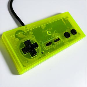 Può includere: Un controller di gioco retrò verde neon con un pad direzionale nero, pulsanti e un cavo nero. La plastica trasparente rivela i circuiti interni. Il controller ha una forma rettangolare ed è progettato per il gioco classico.