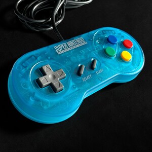Könnte beinhalten: Ein transparent-blauer Super Nintendo Entertainment System-Controller mit einem schwarzen Kabel. Der Controller hat graue Richtungstasten, farbige Aktionstasten und den Text "SUPER NINTENDO ENTERTAINMENT SYSTEM".