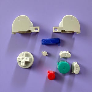 Może przedstawiać: Asortyment przycisków do kontrolera gier w różnych kolorach, w tym białym, niebieskim, czerwonym i zielonym. Elementy są ułożone na fioletowej powierzchni. Przyciski obejmują pady kierunkowe, przyciski barkowe i przyciski funkcyjne.