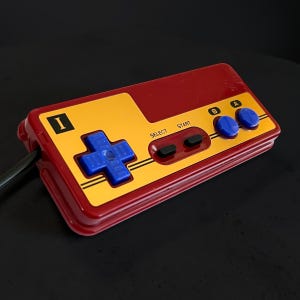 May include: A retro-style video game controller in red and yellow with blue buttons. The controller has a directional pad, select and start buttons, and A and B buttons. The controller is connected to a black cable.