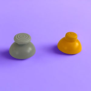 May include: Two colorful joystick thumb grips. One is gray with a circular textured top, and the other is yellow. The grips are set against a solid purple background. These are replacement parts for gaming controllers.