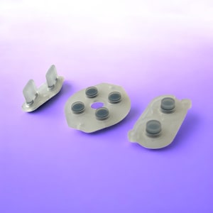 Pode incluir: Três almofadas de botões de silicone cinza translúcido sobre um fundo roxo. Uma almofada tem duas protuberâncias retangulares, outra tem cinco botões circulares e a terceira tem dois botões. Provavelmente são peças de reposição.