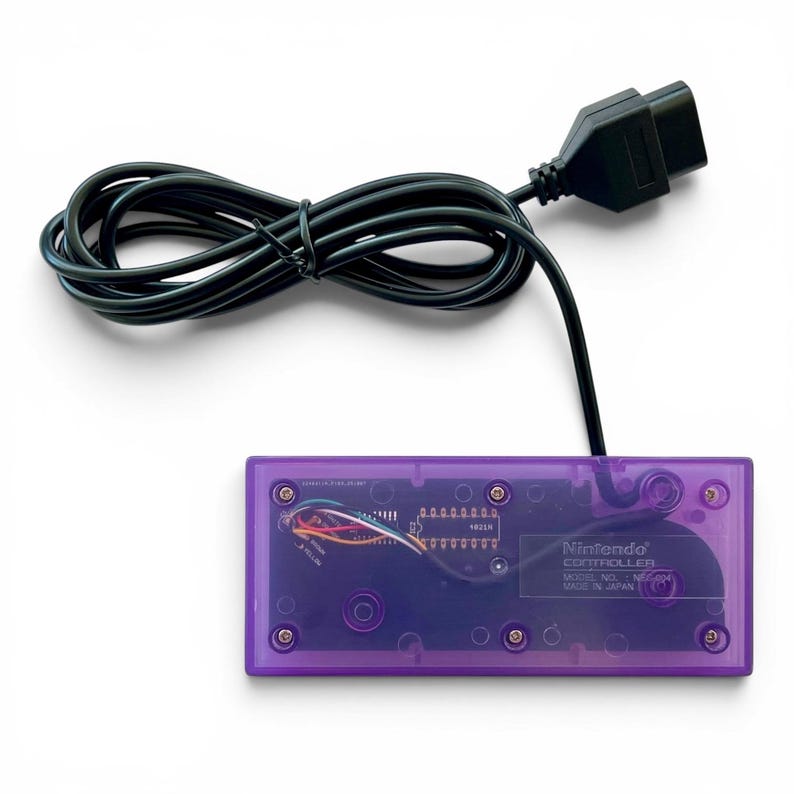 Op de afbeelding: Een transparante paarse Nintendo-controller met een zwarte kabel en connector. De interne componenten van de controller zijn zichtbaar. De tekst "Nintendo Controller" en "Made in Japan" staan op de controller.
