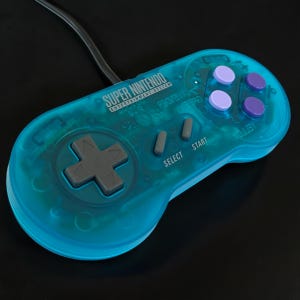 May include: A translucent teal Super Nintendo Entertainment System game controller featuring a grey directional pad, two grey Select and Start buttons, and four action buttons in alternating shades of light and dark purple with a black cord.
