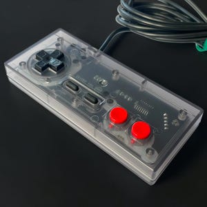May include: A rectangular video game controller featuring a transparent plastic casing that reveals internal electronic components. It includes a black directional pad, two black oval buttons, and two bright red circular action buttons.