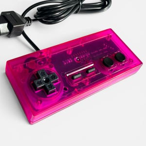 Pode incluir: Um controle de videogame rosa transparente com um direcional preto, botões e um cabo preto. O controle tem formato retangular e design retrô, com componentes internos visíveis.