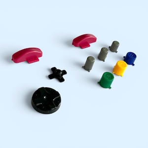Könnte beinhalten: Eine Sammlung farbiger Videospiel-Controller-Tasten und Richtungspads. Die Tasten sind in verschiedenen Farben gehalten, darunter Rot, Grün, Gelb, Blau und Grau. Das Richtungspad ist schwarz. Die Knöpfe sind auf einer hellblauen Oberfläche angeordnet.