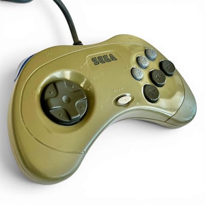 May include: A gold-colored Sega Genesis controller with a directional pad, gray buttons, and a start button. The word "SEGA" is printed on the controller. A black cable extends from the top of the controller.