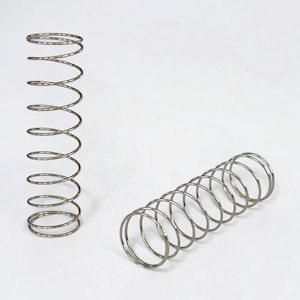 May include: Two silver-colored coil springs are displayed against a white background. One spring stands upright, while the other lies horizontally. The springs are made of metal and have a spiral design. They are likely used for mechanical purposes.