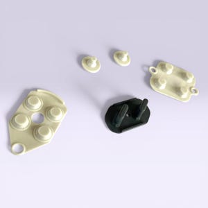 May include: An assortment of off-white and dark green rubber components, likely for electronics or a similar device. The pieces include button-like shapes, a rectangular pad with buttons, and a curved piece with multiple circular protrusions.
