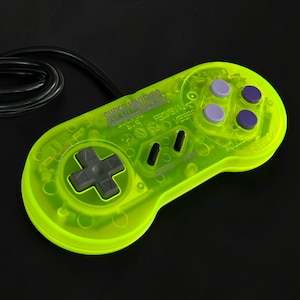 Puede incluir: Un mando de videojuegos translúcido de color amarillo neón con un cable negro. El dispositivo cuenta con una cruceta gris, dos botones ovalados negros etiquetados como Select y Start, y cuatro botones de acción circulares en tonos morados.