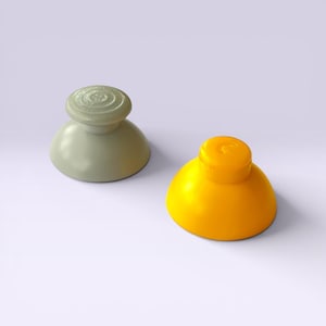 May include: Two colorful joystick thumb grips. One is a pale green, the other is bright yellow. Both have a rounded base and a circular top with a textured design. The grips are designed for video game controllers.