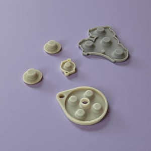 Może przedstawiać: Asortyment jasnoszarych i kremowobiałych gumowych przycisków i komponentów, prawdopodobnie do elektroniki. Elementy ułożone są na jednolitym fioletowym tle. Kształty obejmują okrągłe przyciski i większe, nieregularne elementy.