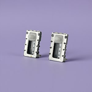 Op de afbeelding: Twee kleine, rechthoekige, zilverkleurige elektronische componenten met een wit plastic stuk aan de bovenkant en een reeks kleine zwarte stippen langs de zijkanten, tegen een effen paarse achtergrond.