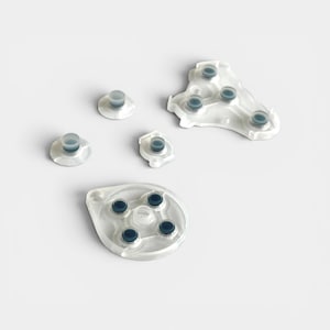 May include: A collection of clear, translucent silicone game controller button pads and individual buttons. The buttons are a light blue-grey color. The pads are various shapes, including round and triangular, with multiple button placements.