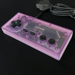 May include: A rectangular video game controller featuring a translucent purple plastic shell. The device includes a black directional pad, two small black rectangular buttons, and two round black action buttons. A black wired cable is attached.