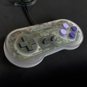 May include: A transparent Super Nintendo Entertainment System game controller with a black wired cord. The controller features a dark gray directional pad, two dark gray center buttons labeled Select and Start, and four purple action buttons.