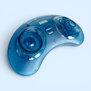 Könnte beinhalten: Ein durchscheinender blauer Sega Saturn Controller. Der Controller hat ein einzigartiges, geschwungenes Design mit einem Steuerkreuz, einer Starttaste und sechs Aktionstasten. Das Wort "SEGA" ist auf der Oberseite aufgedruckt.