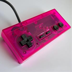 May include: A translucent pink retro games controller with a black directional pad, buttons, and a black cable. The internal circuitry is visible through the transparent plastic.