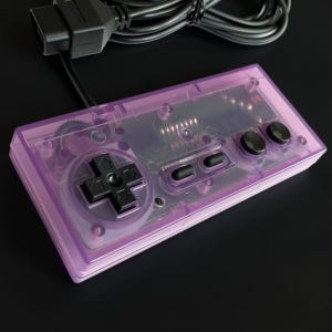 May include: A rectangular video game controller featuring a translucent purple plastic shell. The device includes a black directional pad, two black oval-shaped buttons, two black circular buttons, and a black wired cable with a connector plug.