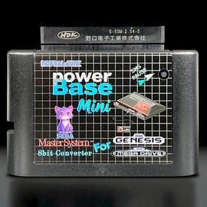 Puede incluir: Convertidor Power Base Mini de 8 bits negro para consolas Sega Master System y Genesis. El cartucho presenta el texto "Power Base Mini", un gráfico de gato morado y el logotipo de Sega.