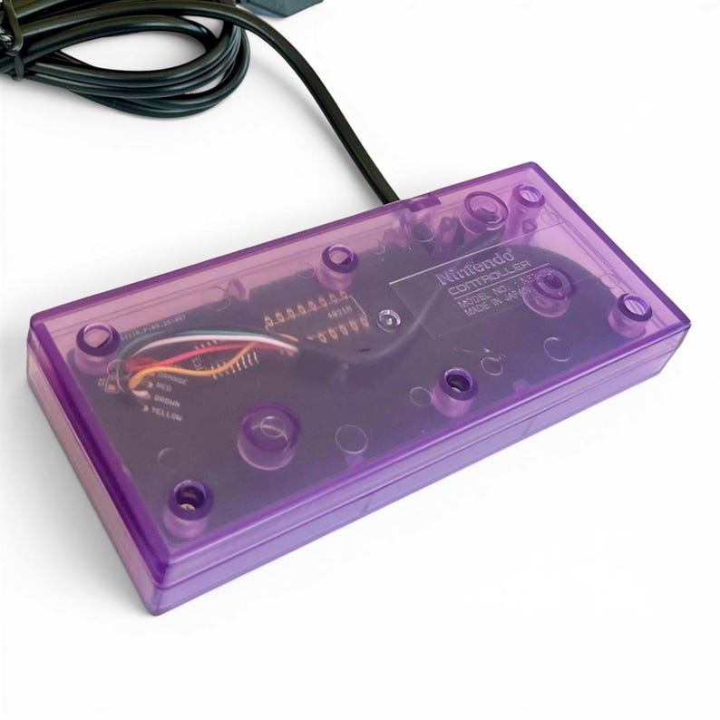 Op de afbeelding: Een transparante paarse Nintendo-controller met een zwarte kabel. De interne componenten van de controller zijn zichtbaar door het doorschijnende plastic. De tekst "Nintendo CONTROLLER" en "MODEL NO. HVC-005" zijn afgedrukt.