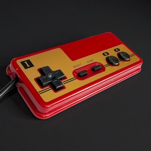 May include: A retro-style red and gold video game controller with a black directional pad, buttons, and a cable. The controller has the letters "I", "A", and "B" on it, along with "SELECT" and "START" buttons.