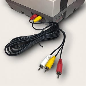 Pode incluir: Uma consola Nintendo Entertainment System (NES) cinza e preta com cabos RCA vermelhos, amarelos e brancos. Os cabos estão ligados à consola e estendem-se para baixo, com os conectores vermelho, amarelo e branco visíveis.